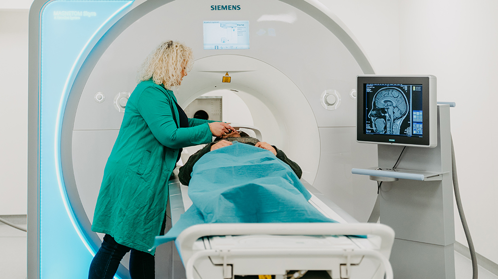 /unimagdeburg_media/Presse/Bilder/Pressemitteilungen/2024/Ein+Patient+liegt+in+einem+MRT_+eine+Frau+legt+ihm+eine+Halterung+f%C3%BCr+den+Kopf+an_+rechts+sieht+man+eine+erste+Bildgebung+eines+menschlichen+Kopfes+%28c%29+Jana+D%C3%BCnnhaupt+Uni+Magdeburg-height-560-width-1000-p-111934.jpg
