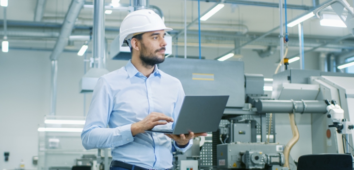 Mann steht mit Laptop in Produktionshalle und prüft die Abläufe (c) Shutterstock / Gorodenkoff